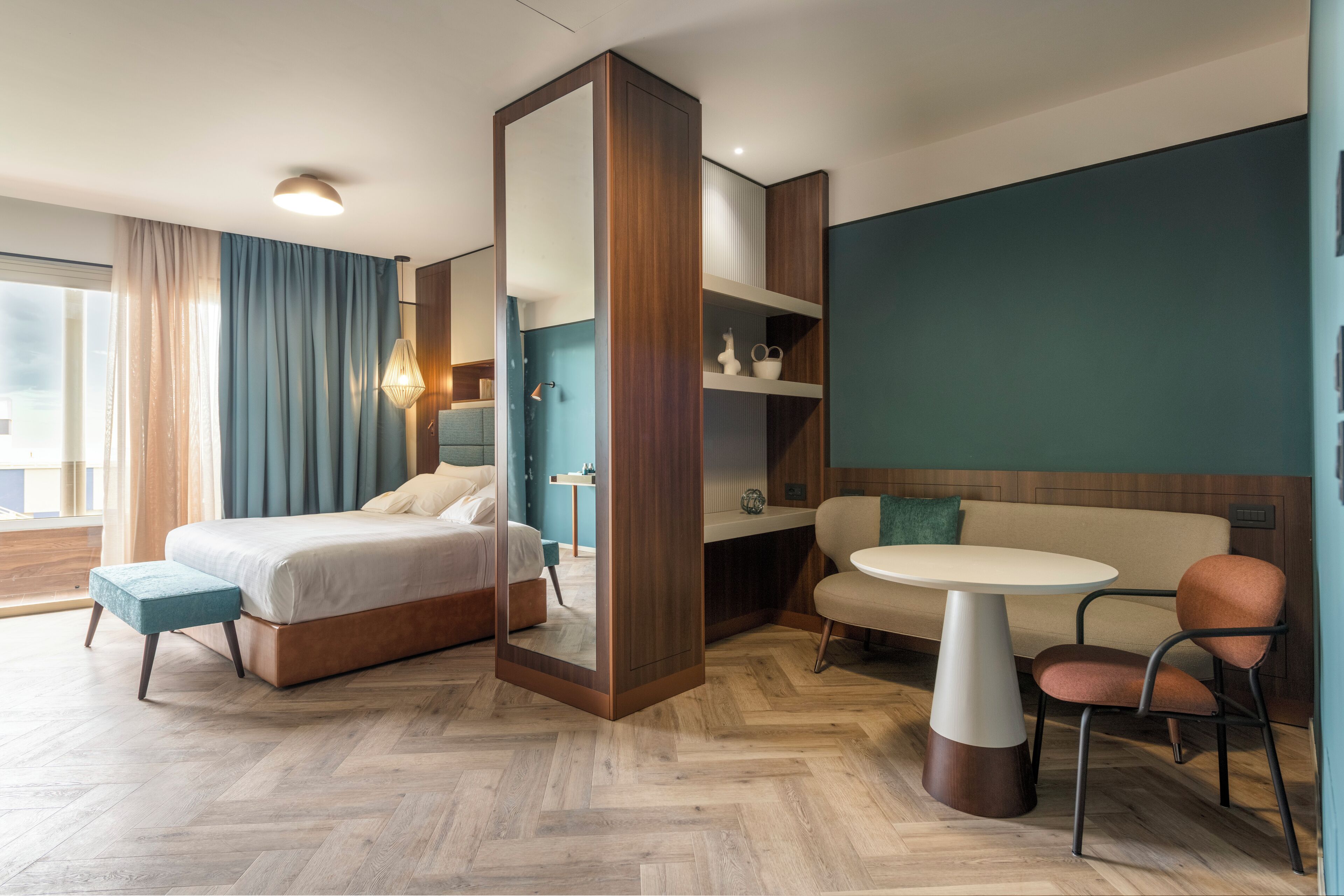 A modern hotel room with a large bed, a sofa, a dining table, and a mirror.