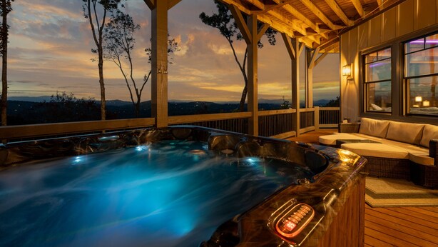 A hot tub area with a wooden deck, a cushioned seating area, and a view of the landscape. - Wine Down Ridge- Blue Ridge GA (Blue Ridge)