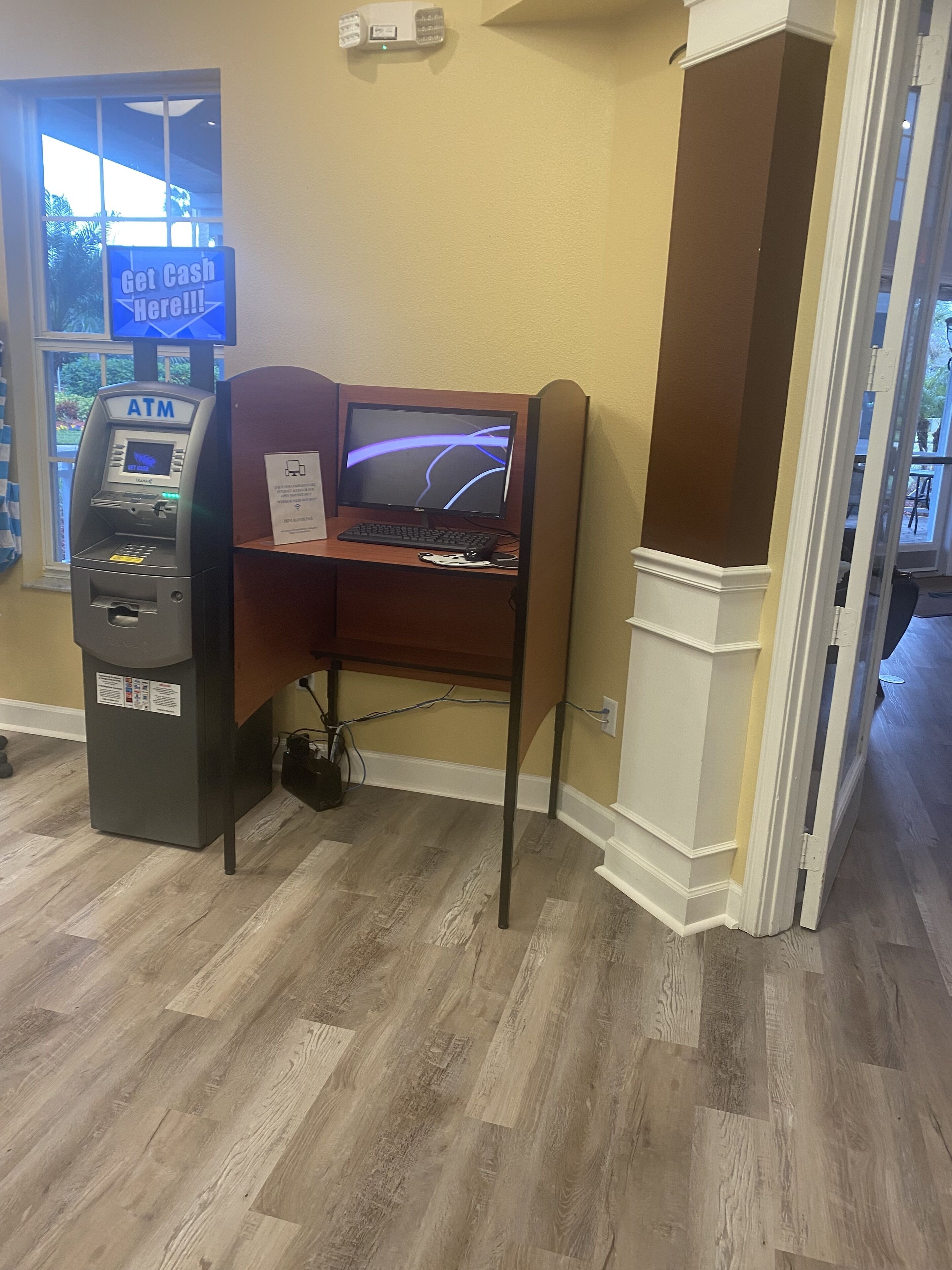 An ATM, a computer desk, and a wooden partition in a room with wooden flooring and a window.