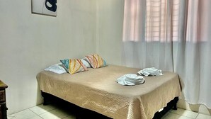 Ein ordentlich bezogenes Bett mit beiger Bettdecke und Kissen, ein kleiner Nachttisch mit Lampe sowie ein Fenster mit transparenten Vorhängen.