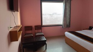 A bedroom with a bed, two chairs, a small table, a TV mounted on the wall, and a window with curtains. - The Kapji Palace (Jaipur)