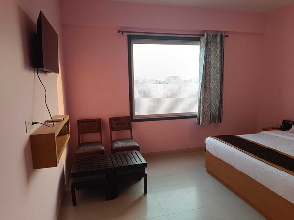 A bedroom with a bed, two chairs, a small table, a TV mounted on the wall, and a window with curtains. - The Kapji Palace (Jaipur)