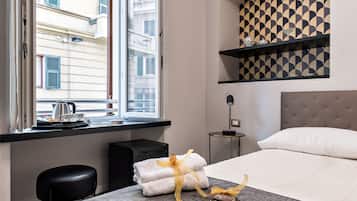 Una camera d'albergo con un letto, una finestra che si affaccia su edifici, un comodino con una lampada e un muro con motivo geometrico.