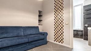 Een moderne woonkamer met een blauwe bank, een wand met geometrisch patroon, en een badkamer zichtbaar door een open deur.