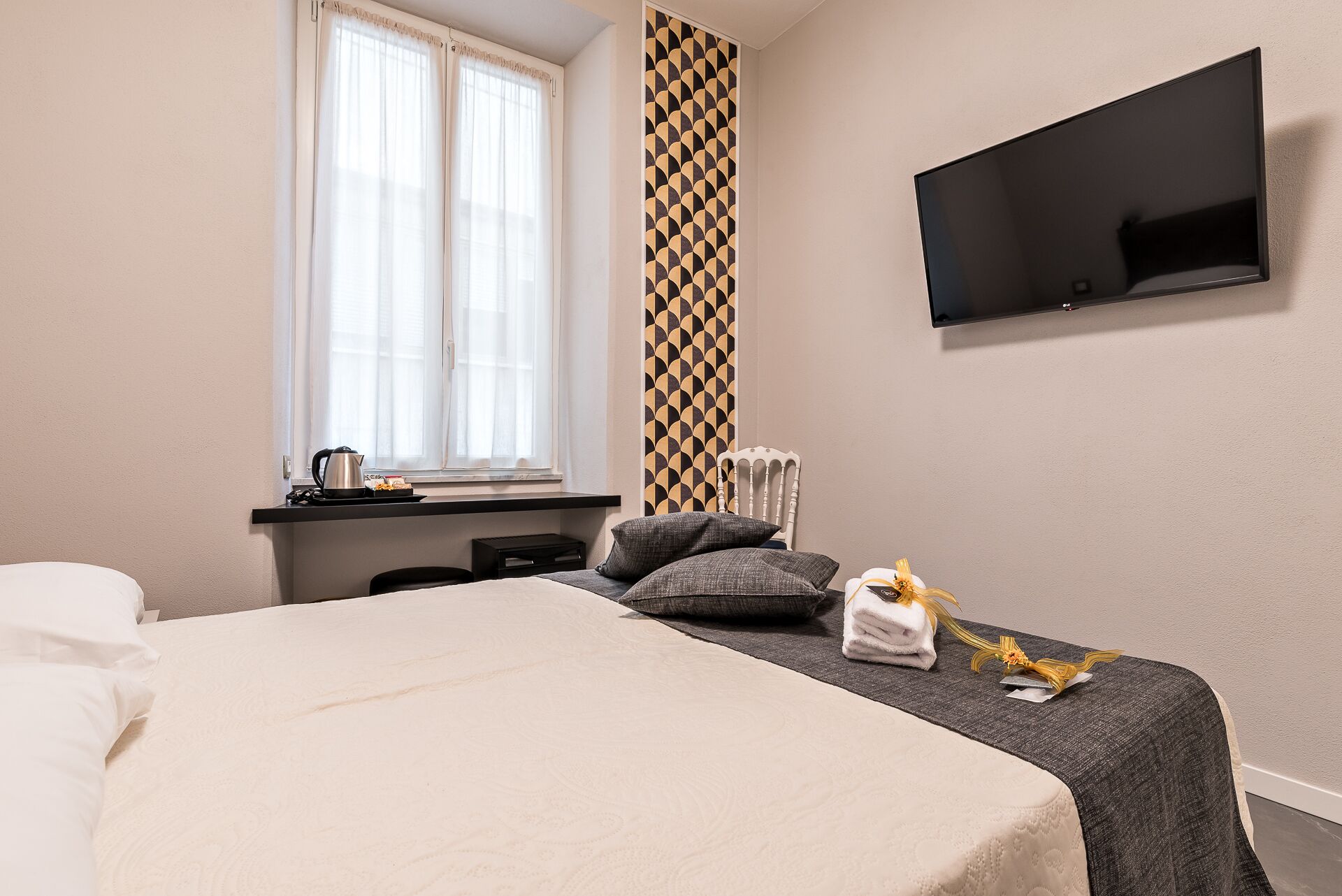 Ein Hotelzimmer mit Bett, Wandfernseher, Fenster mit Vorhängen, Heizkörper und kleinem Tisch mit Wasserkocher und Tablett.