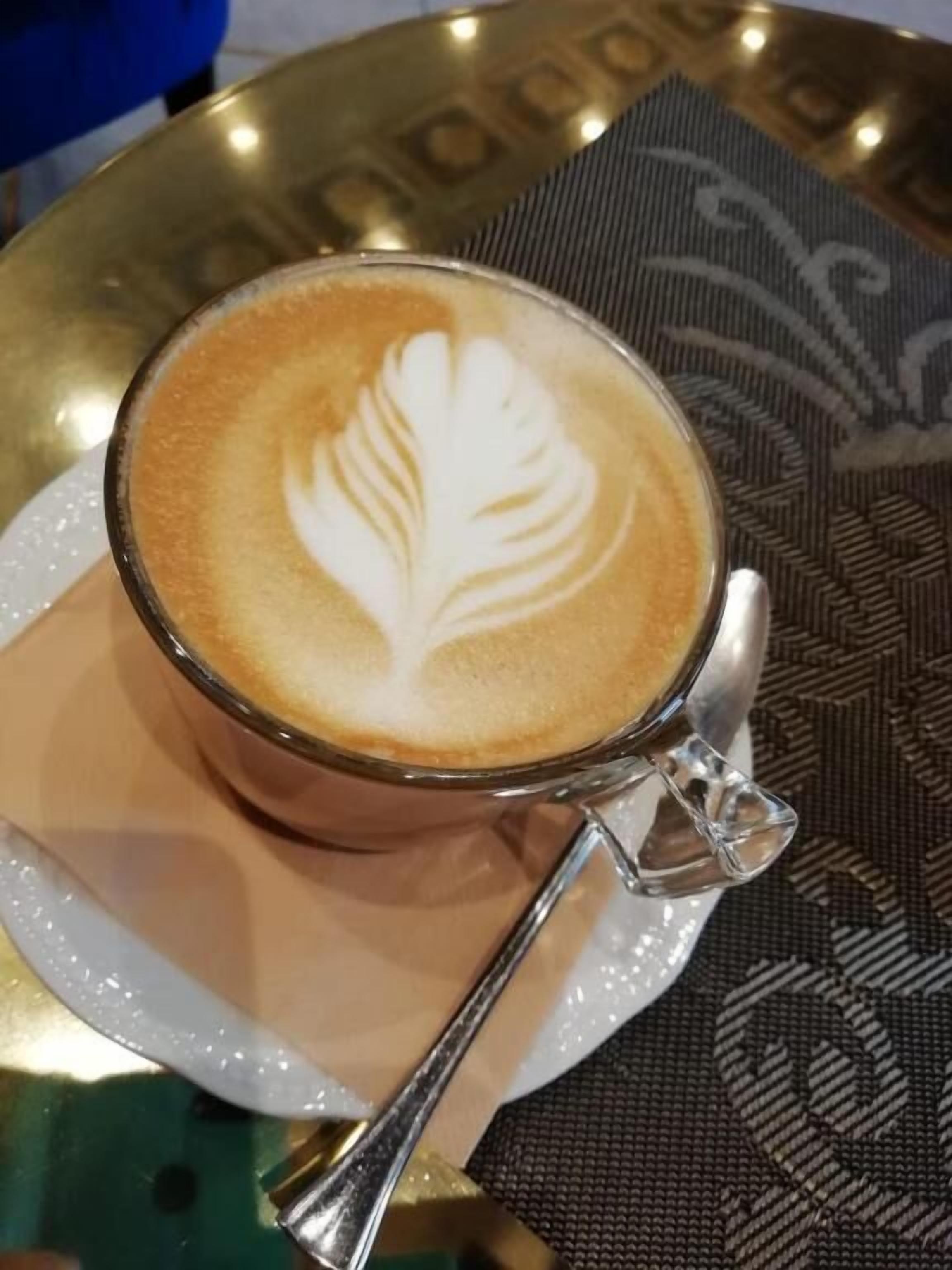 Une tasse de café ornée d’un motif en latte art, posée sur une soucoupe, le tout placé sur une table recouverte d’une nappe à motifs.