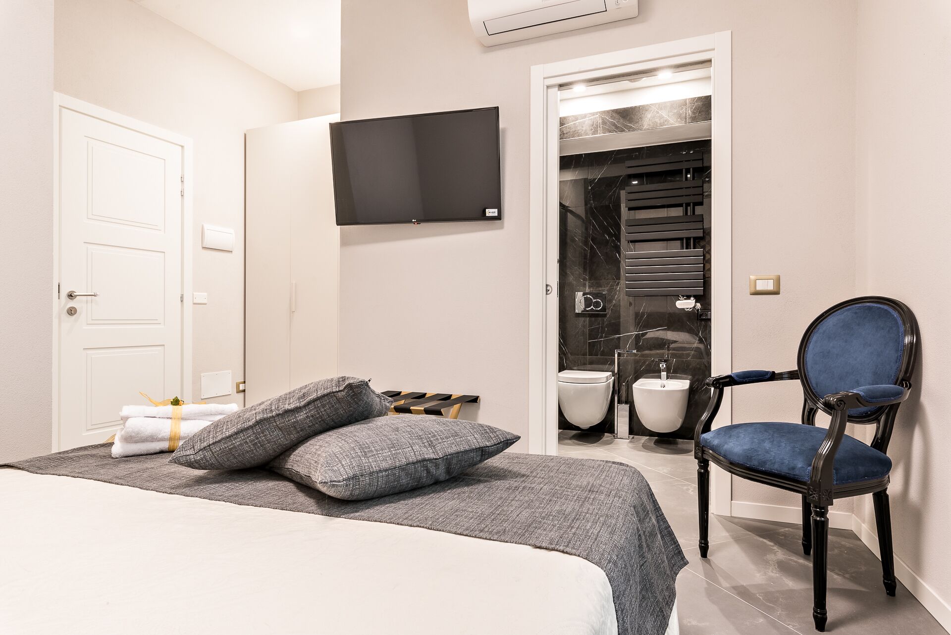 Hotellihuone, jossa on sänky, tuoli, televisio ja kylpyhuone, jotka näkyvät avoimesta ovesta.