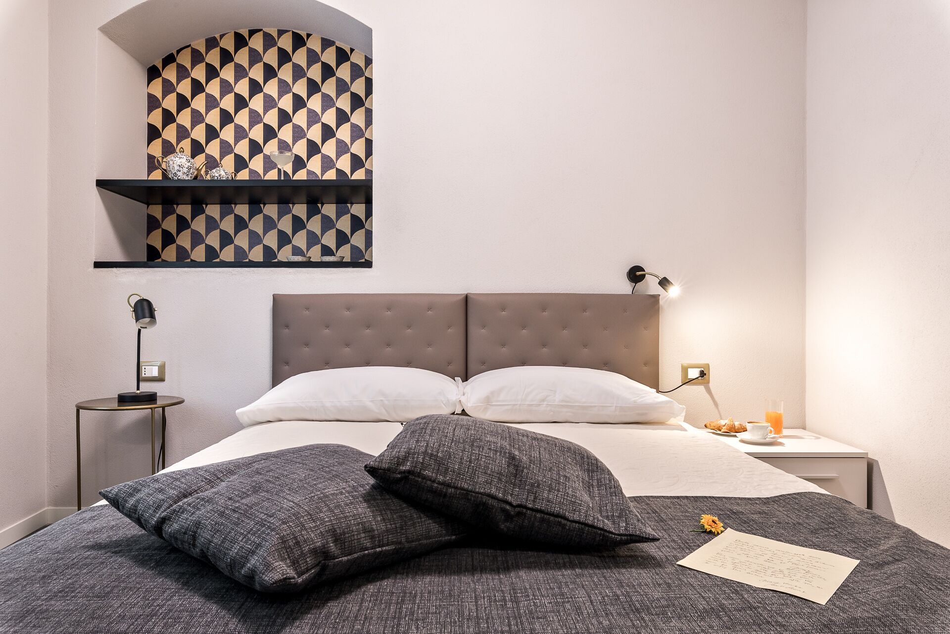 Un dormitorio con cabecera capitonada, dos almohadas blancas, una manta gris y una mesita de noche con una lámpara y un vaso de jugo de naranja.