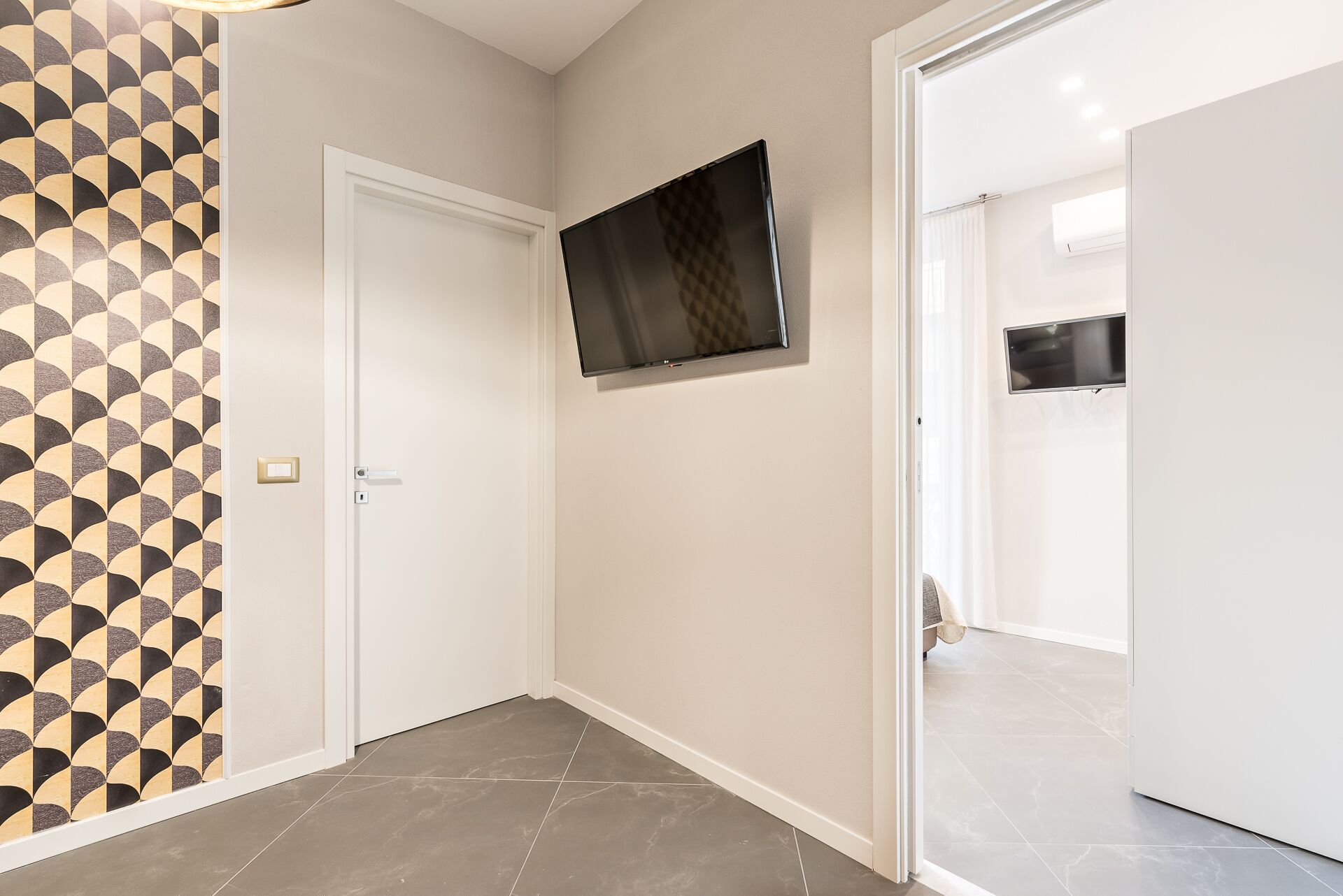 A hallway with a tiled floor, a door with a geometric patterned wallpaper, and a mounted flat-screen TV.