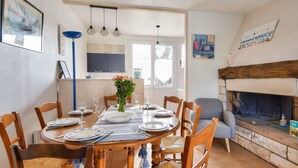 Une salle à manger avec une table en bois dressée pour six personnes, entourée de chaises en bois. Une cheminée est visible sur la droite, et un coin cuisine se trouve au fond de la pièce.
