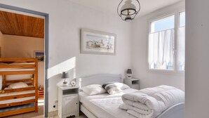 Une chambre avec un lit superposé, un lit simple, une table de chevet avec une lampe, un tableau encadré au mur et une fenêtre avec des rideaux.