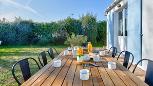 Une table de salle à manger en bois, mise avec des tasses, une bouteille et des couverts, entourée de chaises, avec un jardin et un bâtiment en arrière-plan.