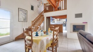A dining area with a table set for four, wooden chairs, a staircase, a kitchen in the background, and a fireplace. - Les Portes en Ré, house with garden for 7 (Les Portes-en-Ré)