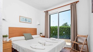 A bedroom with a large bed, a wooden headboard, a nightstand with a lamp, and a view of greenery through a sliding door. - Bini Rosa - Five Bedroom Villa, Sleeps 10 (Binibeca)
