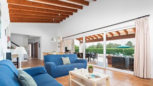 A spacious living room with a blue sofa, a coffee table, and a large sliding glass door leading to an outdoor patio. - Bini Rosa - Five Bedroom Villa, Sleeps 10 (Binibeca)