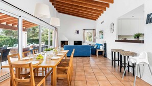 A spacious dining area with a wooden table and chairs, a blue sofa, and a kitchen area with a clock and cabinets. - Bini Rosa - Five Bedroom Villa, Sleeps 10 (Binibeca)
