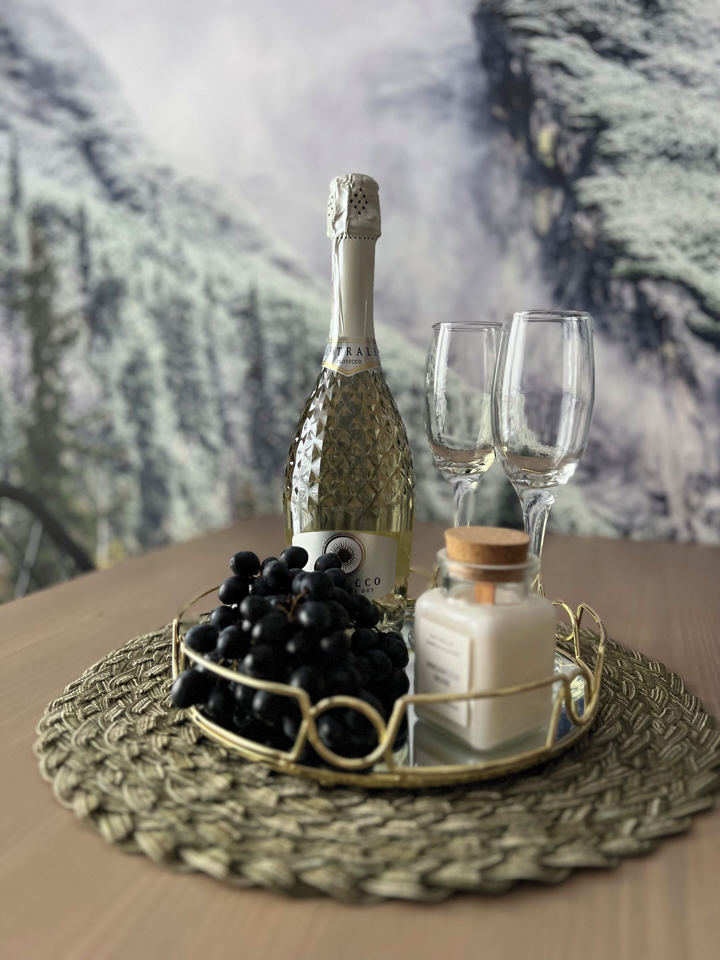 Una botella de champán, dos copas, un cuenco de uvas y una vela sobre una bandeja de mimbre.