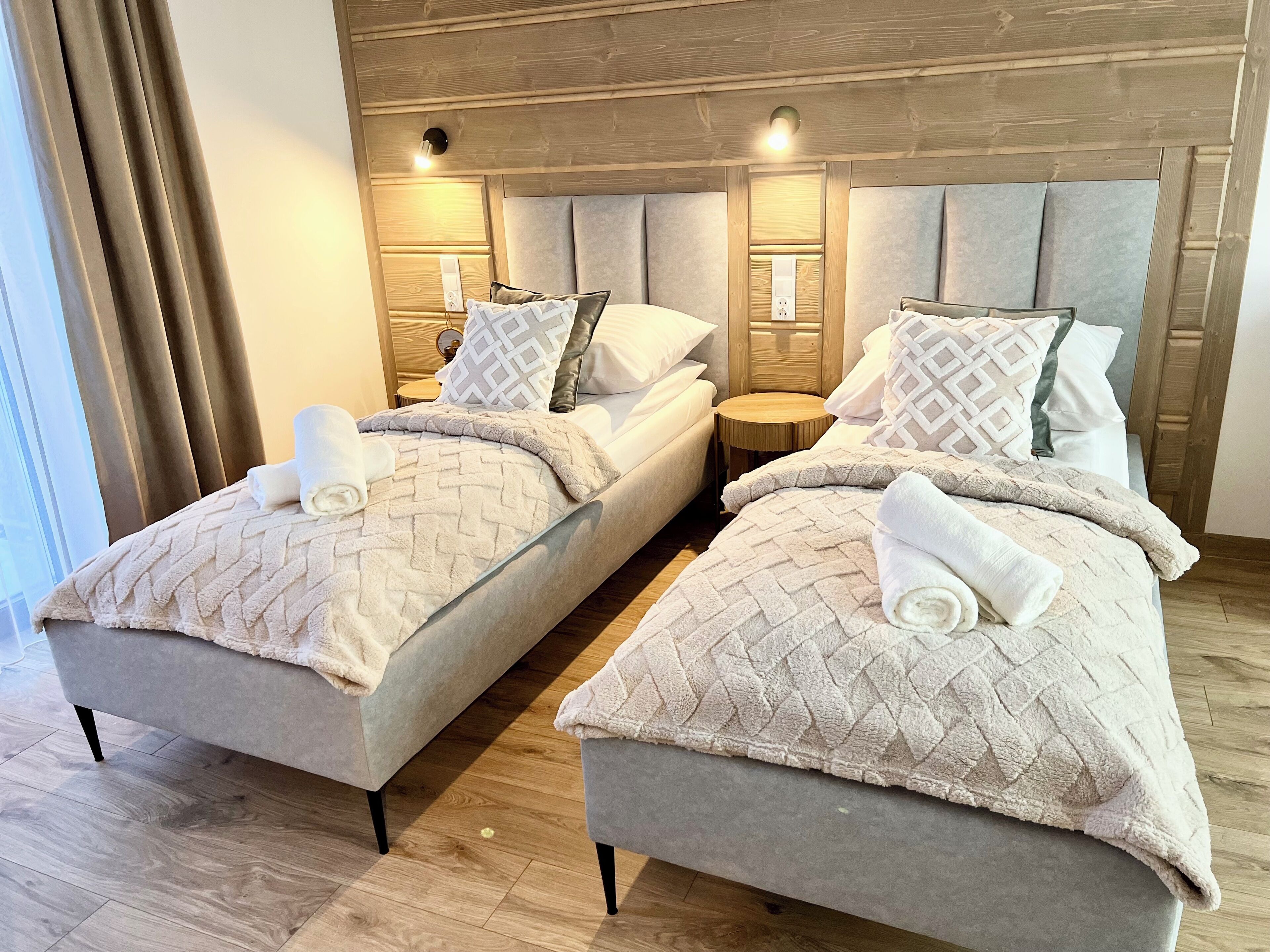 A hotel room with two beds, each with a headboard, a small round table, and a window with curtains.