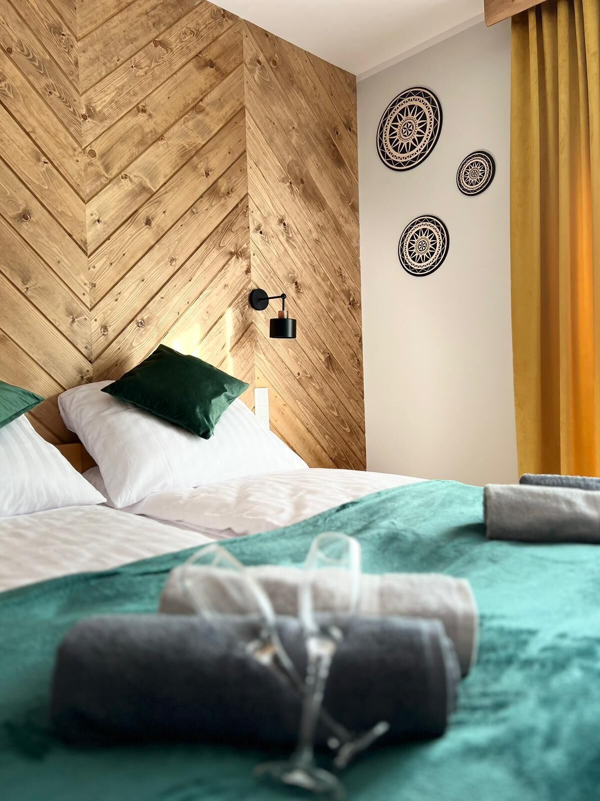 A neatly made bed with green and white bedding, a wooden wall with a chevron pattern, and a wall-mounted lamp.