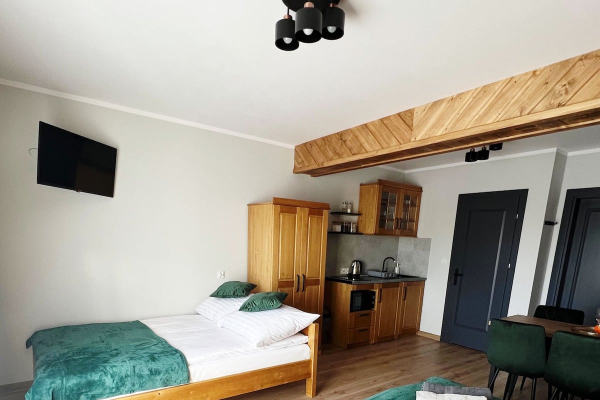 A modern bedroom with two beds, a wooden wardrobe, a kitchenette, and a TV mounted on the wall.