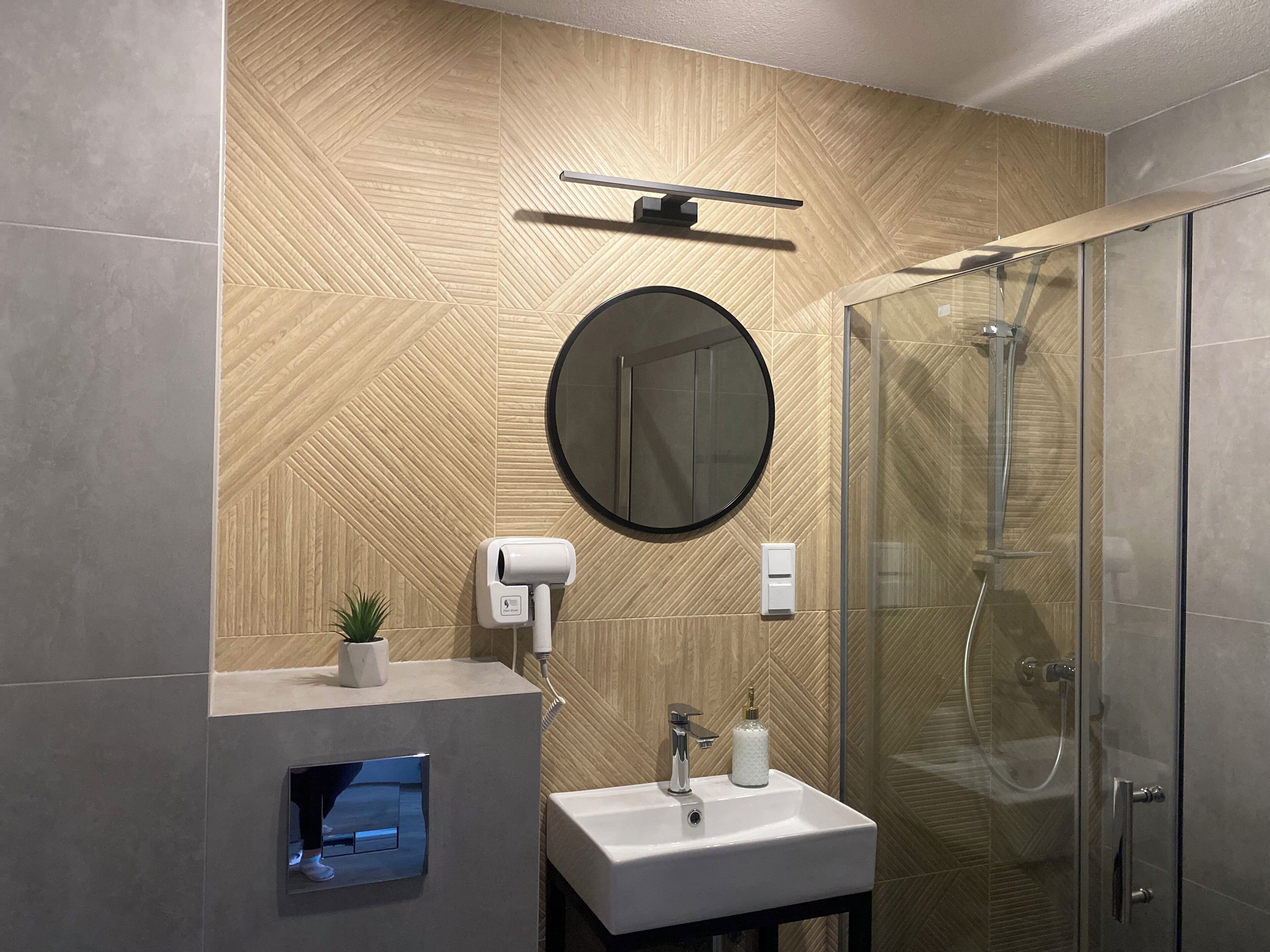 Un baño moderno con patrón de pared de madera en espiga, un espejo redondo, ducha con cerramiento de vidrio y lavabo empotrado con una pequeña planta en maceta.