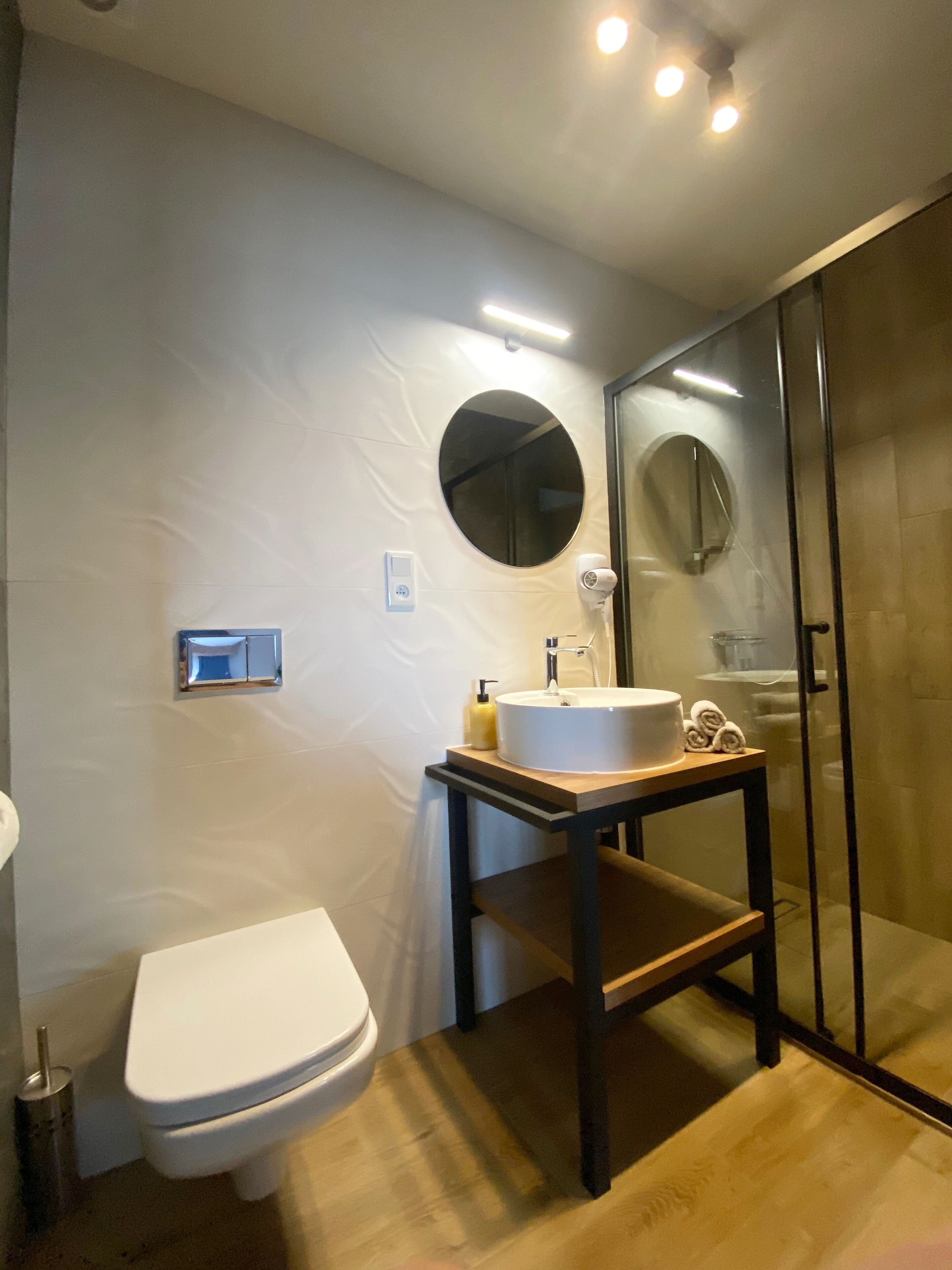 Une salle de bain moderne avec des toilettes blanches, un miroir rond, un lavabo doté d’un plan de travail en bois et une cabine de douche en verre.