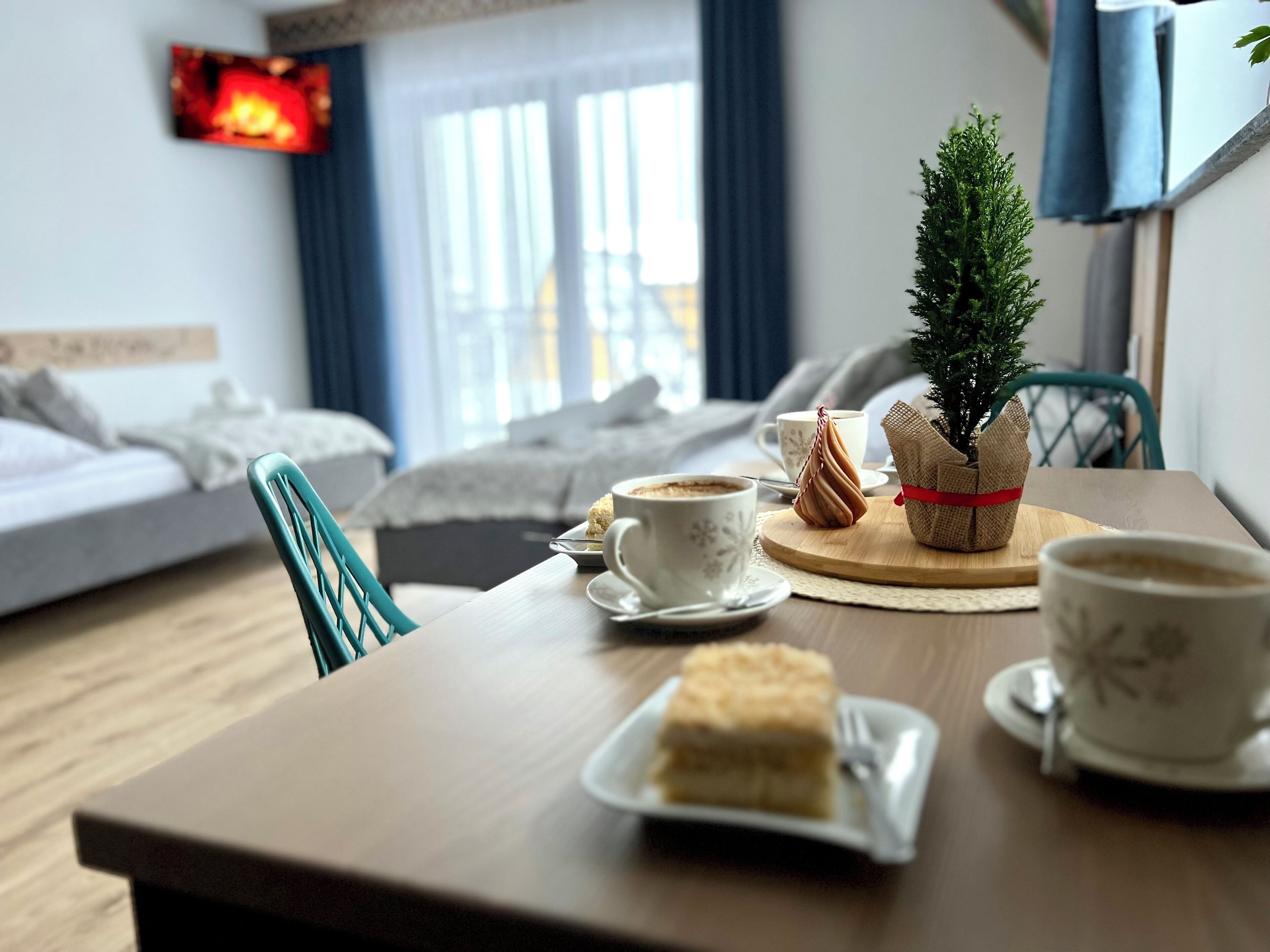 A hotel room with a dining table set with a slice of cake, a cup of coffee, and a small potted plant.