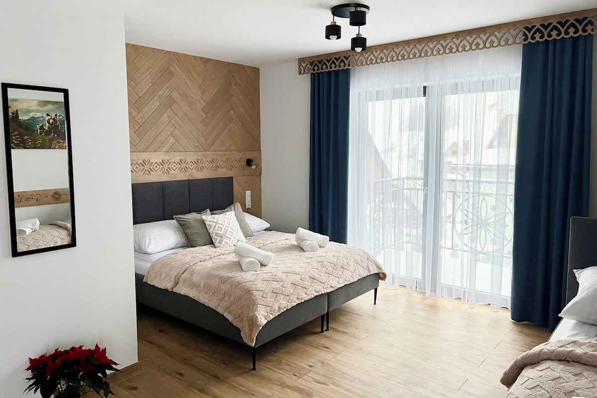 A modern bedroom with a herringbone-patterned headboard, a bed with a beige quilt, and a large window with blue curtains.
