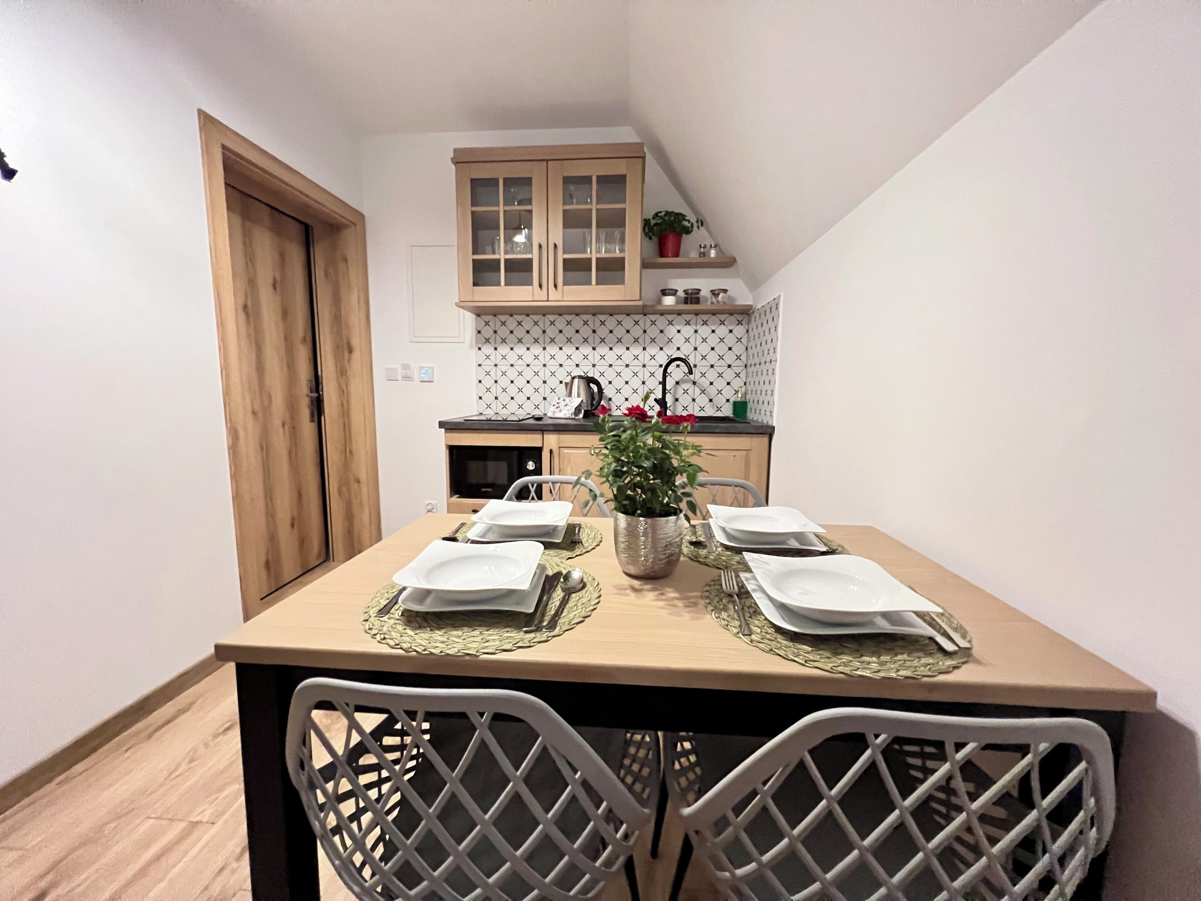 Un pequeño comedor con una mesa de madera dispuesta para cuatro, rodeada de sillas. Hay una planta en maceta sobre la mesa, y se observa una cocina al fondo con armarios y un lavabo.
