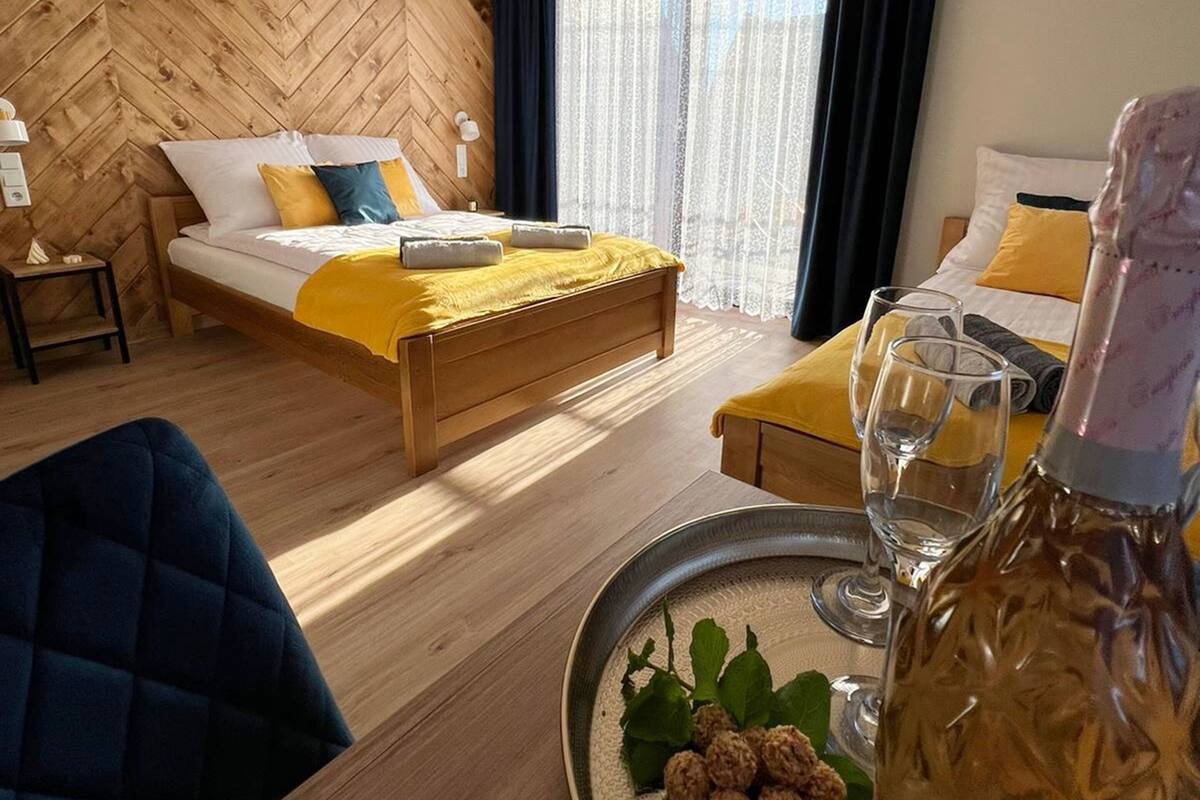 A hotel room with a wooden herringbone wall, a bed with white and yellow bedding, a second bed with yellow and white bedding, a dining table with a tray of nuts and a bottle of champagne, and a blue armchair.