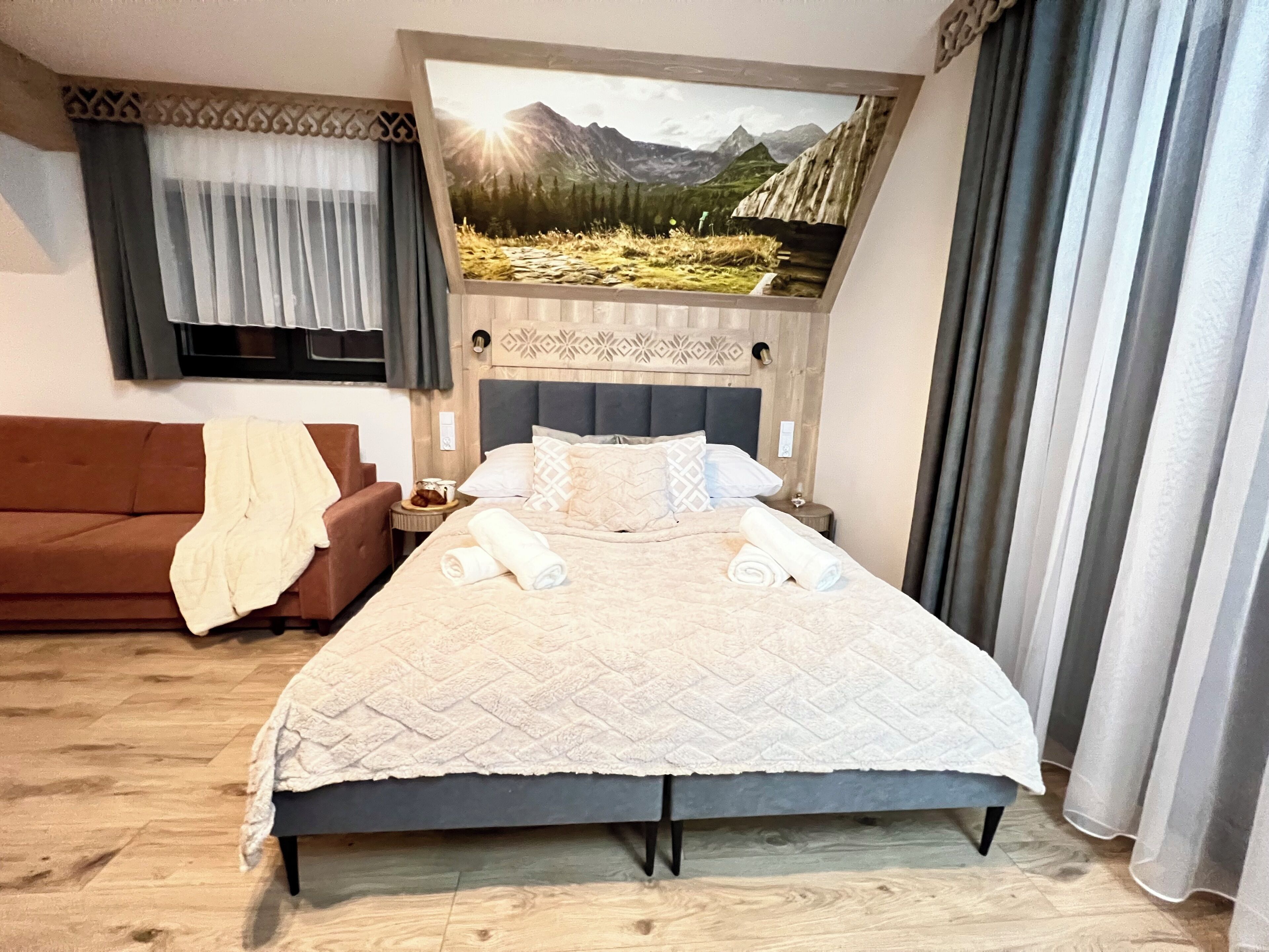 Habitación de hotel con cama, sofá marrón, mesita de noche y un cuadro de un paisaje montañoso.