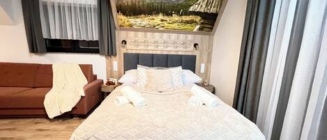 Habitación de hotel con cama, sofá marrón, mesita de noche y un cuadro de un paisaje montañoso.
