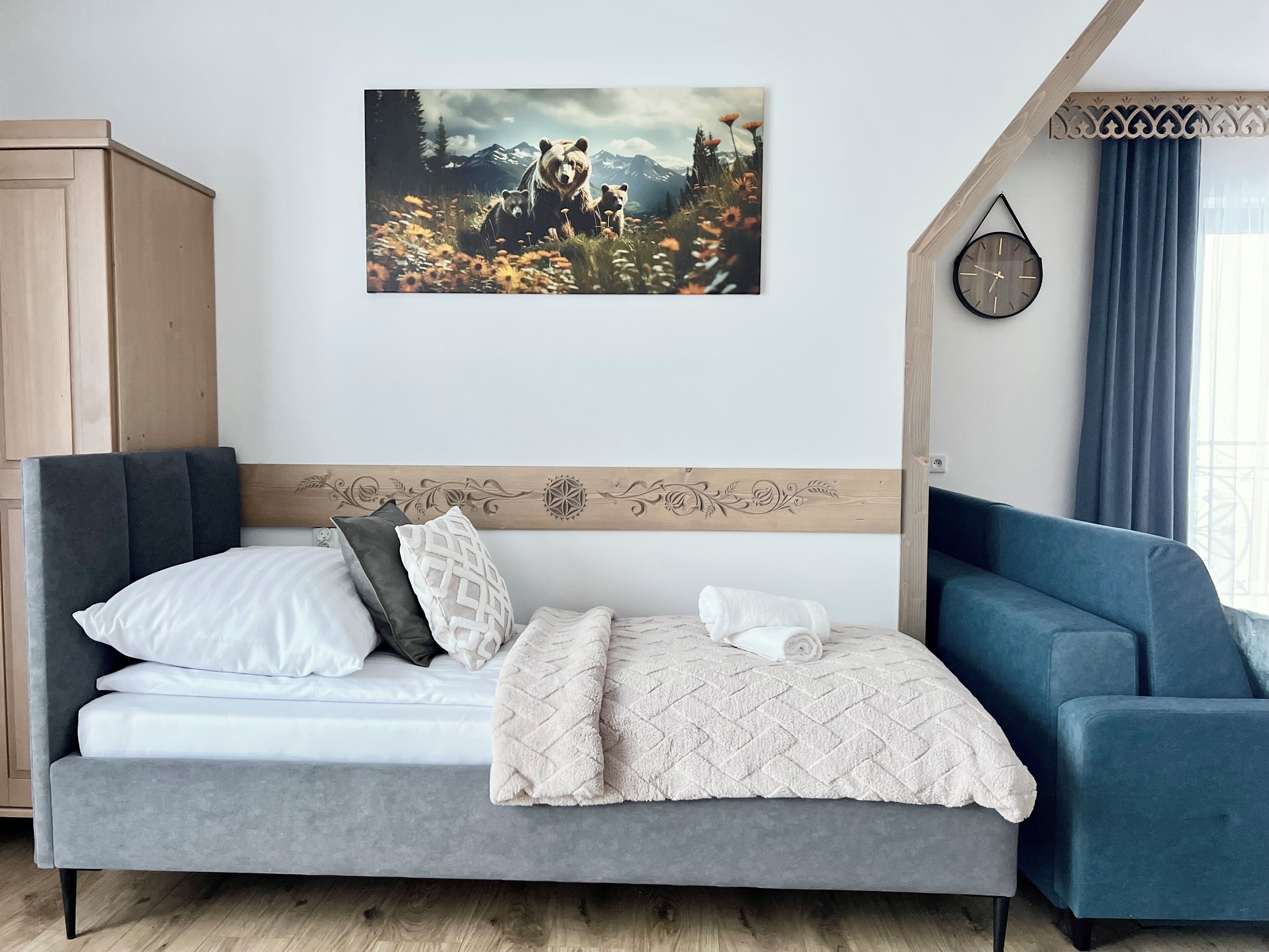 A bedroom with a bed, a blue armchair, a wooden headboard, a wall clock, and a painting of a bear family.