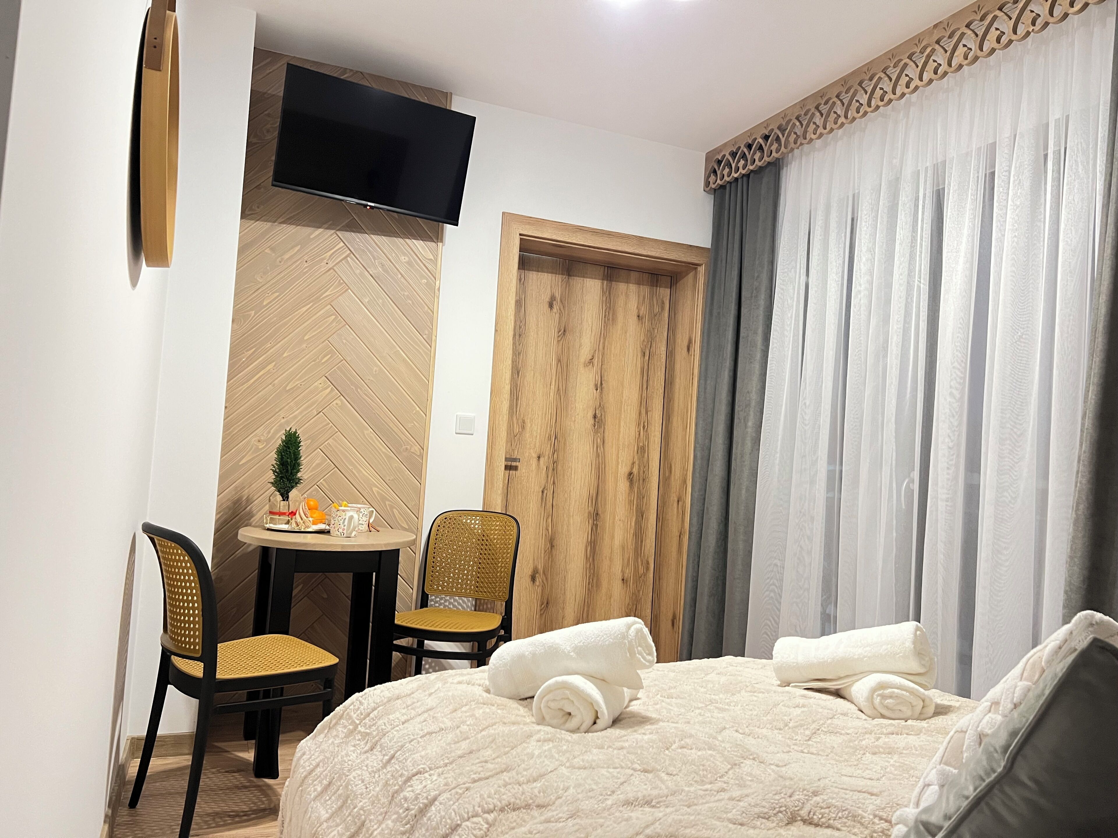 A hotel room with a bed, a small table with chairs, a TV mounted on the wall, and a wooden door.