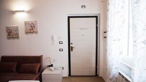 A room with a brown sofa, a small white table with a vase, and a white door with a nameplate. - Appartamento Viale Patrono D'italia (Santa Maria degli Angeli)