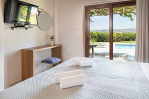 A hotel room with a large bed, a TV mounted on the wall, a small table with a vase, and a view of a swimming pool and greenery outside.