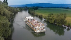 A riverboat with a covered deck and seating area, moving along a river surrounded by greenery and fields. - Boutique Boat Hotel Attila (Bas-Vully)