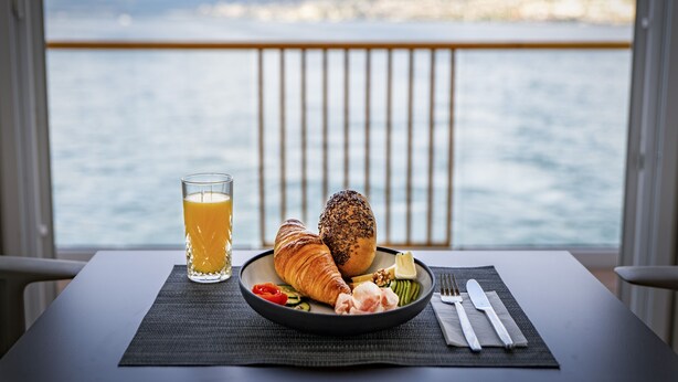 A breakfast setting with croissants, a glass of orange juice, and a plate of vegetables on a table by a window overlooking a body of water. - Boutique Boat Hotel Attila (Bas-Vully)