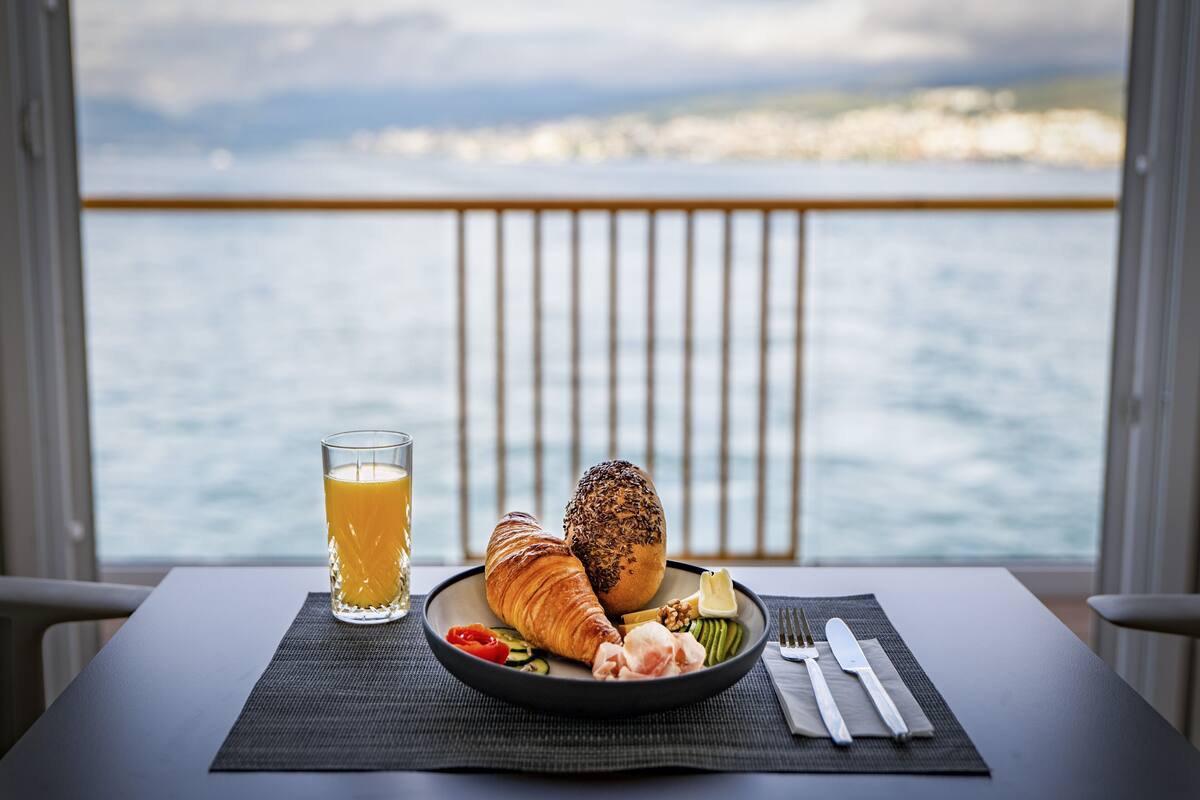 A breakfast setting with croissants, a glass of orange juice, and a plate of vegetables on a table by a window overlooking a body of water.
