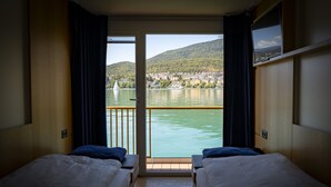 A hotel room with two beds, a balcony with a view of a lake and a town, and a window with a picture frame. - Boutique Boat Hotel Attila (Bas-Vully)