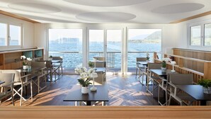 A modern dining area with tables and chairs, large windows with a view of the sea, and a wooden ceiling. - Boutique Boat Hotel Attila (Bas-Vully)