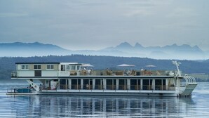A large, multi-deck boat with the name "ATTILA" on the side, docked on calm water with a mountainous backdrop. - Boutique Boat Hotel Attila (Bas-Vully)