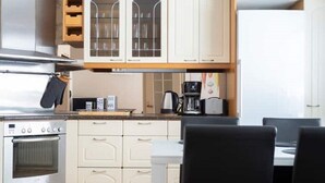 Een kleine keuken met witte kasten, een inbouwoven, een koffiezetapparaat en een eettafel met stoelen.