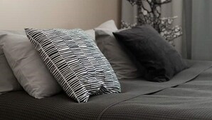 Een bed met grijze beddengoed en twee kussens, één gestreept en één effen, in een kamer met een grijze muur en een raam met een ontwerp van een boom.