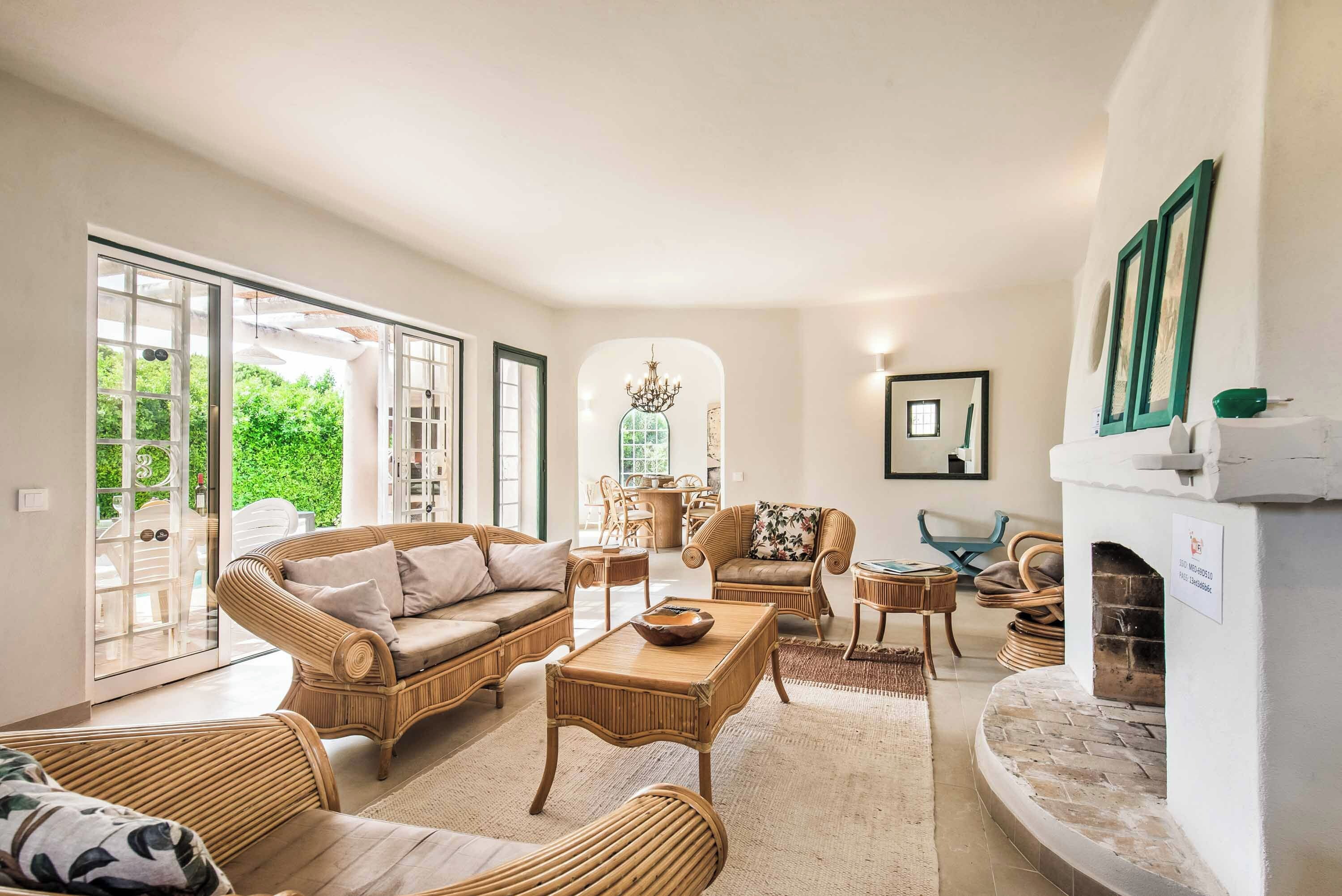 A spacious living room with wicker furniture, a fireplace, and large windows offering a view of greenery.