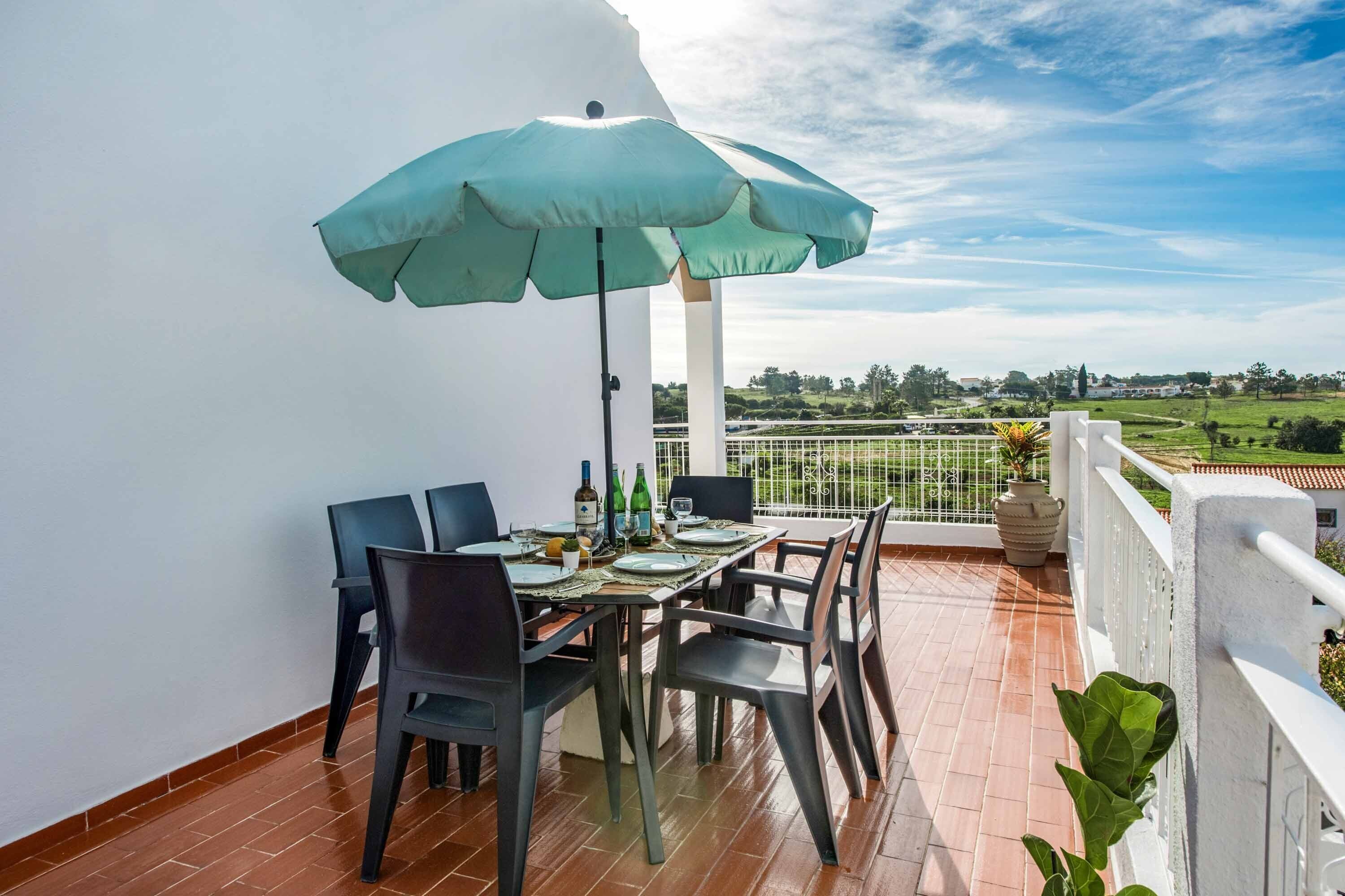 A balcony with a table set for four, a green umbrella, and a view of a green landscape.