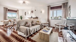 Sala de estar con sofá, sillas, una mesa de centro y televisor. Hay ventanas con cortinas, un comedor y una planta.