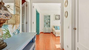 Un couloir avec un mur orné de motifs, un banc bleu et une porte turquoise donnant sur une pièce où se trouvent une commode blanche et une chaise bleue.