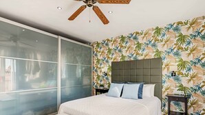 Une chambre à coucher avec un lit, un banc, un ventilateur de plafond et un papier peint aux motifs tropicaux.