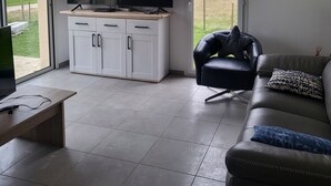 Een moderne woonkamer met een flatscreen televisie, witte kasten, een leren bank en een groot raam met uitzicht op de omgeving.